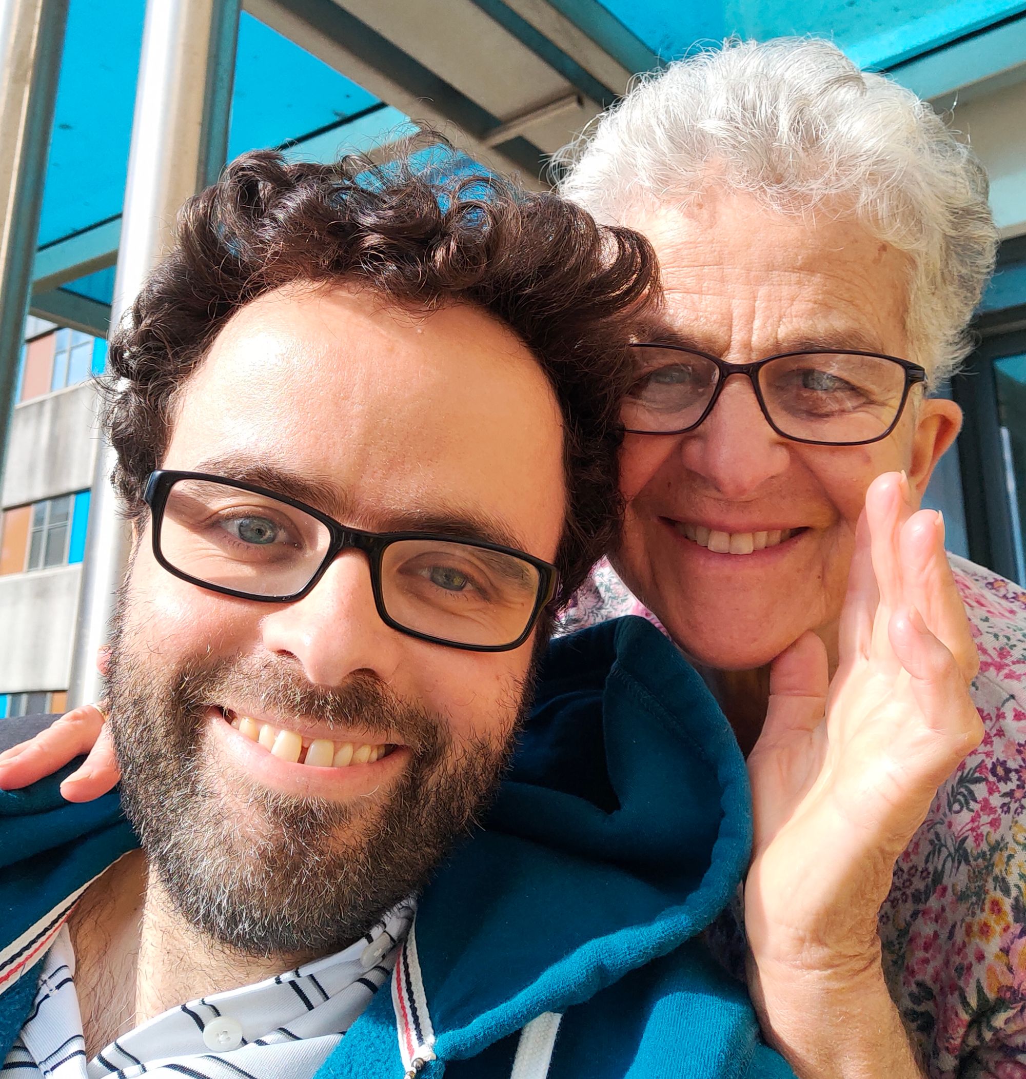 A smiling selfie of Peter and his Mother. They are both white, and share similar eyes, nose and curly hair. Peter has glasses and a beard. Mummy Fremlin has white hair, glasses, one hand on his shoulder and the other held vertically as a hello. 