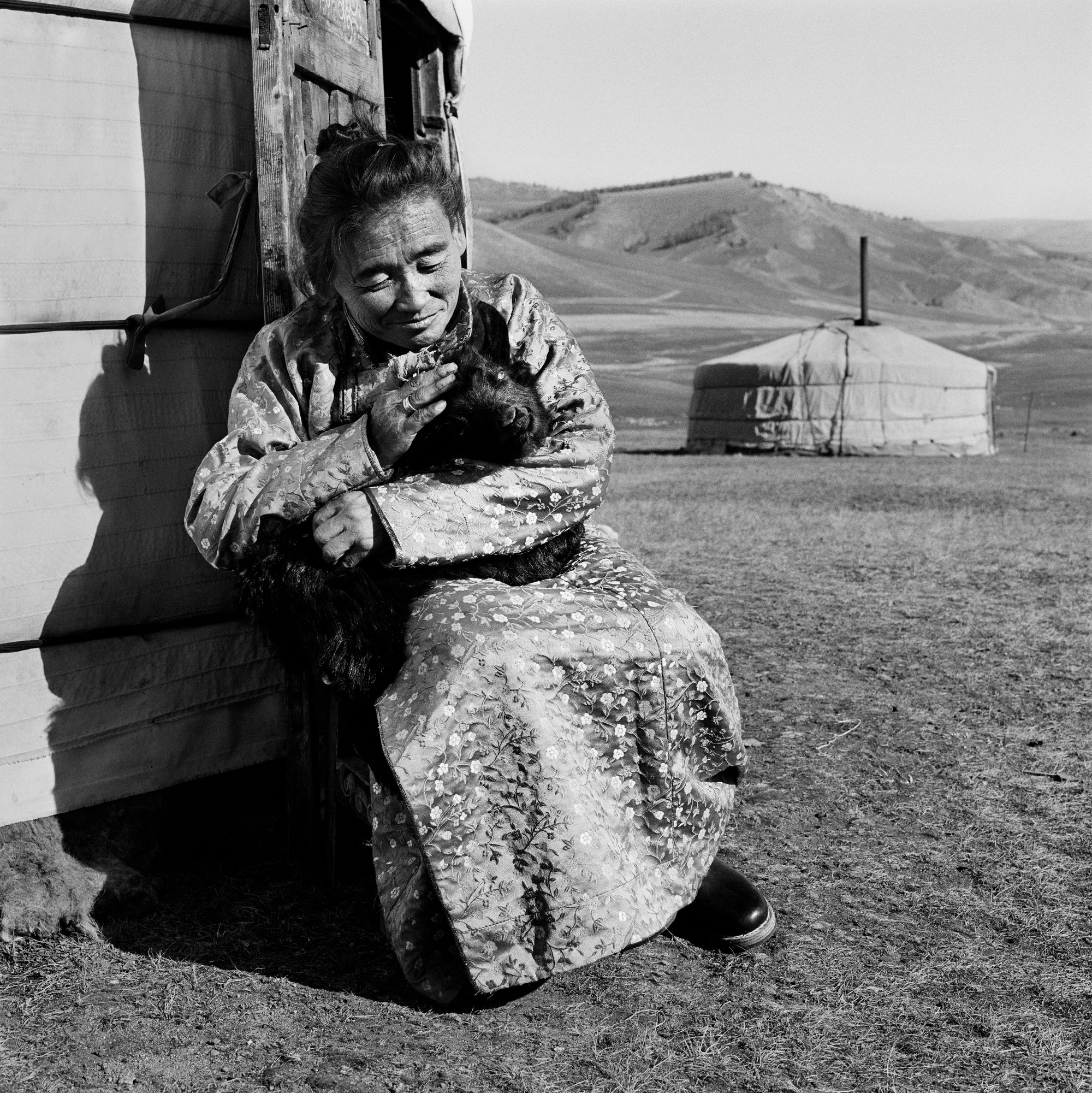 Black and white photo of an older woman, looking tenderly at the small goat in her hands. She wears a body-length jacket patterned with flowers. She sits by a yurt, with another visible in the background, hills stretching behind it.