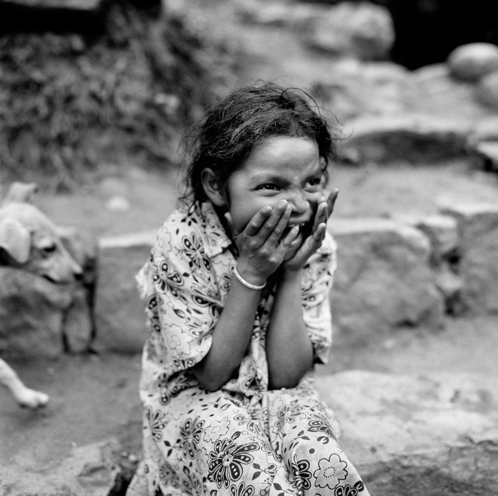 Black and white photo of a young girl wide-mouthed in laughter and joy, hands to her face, wearing a dress. She is seated outside, with a dog walking into the frame behind her.