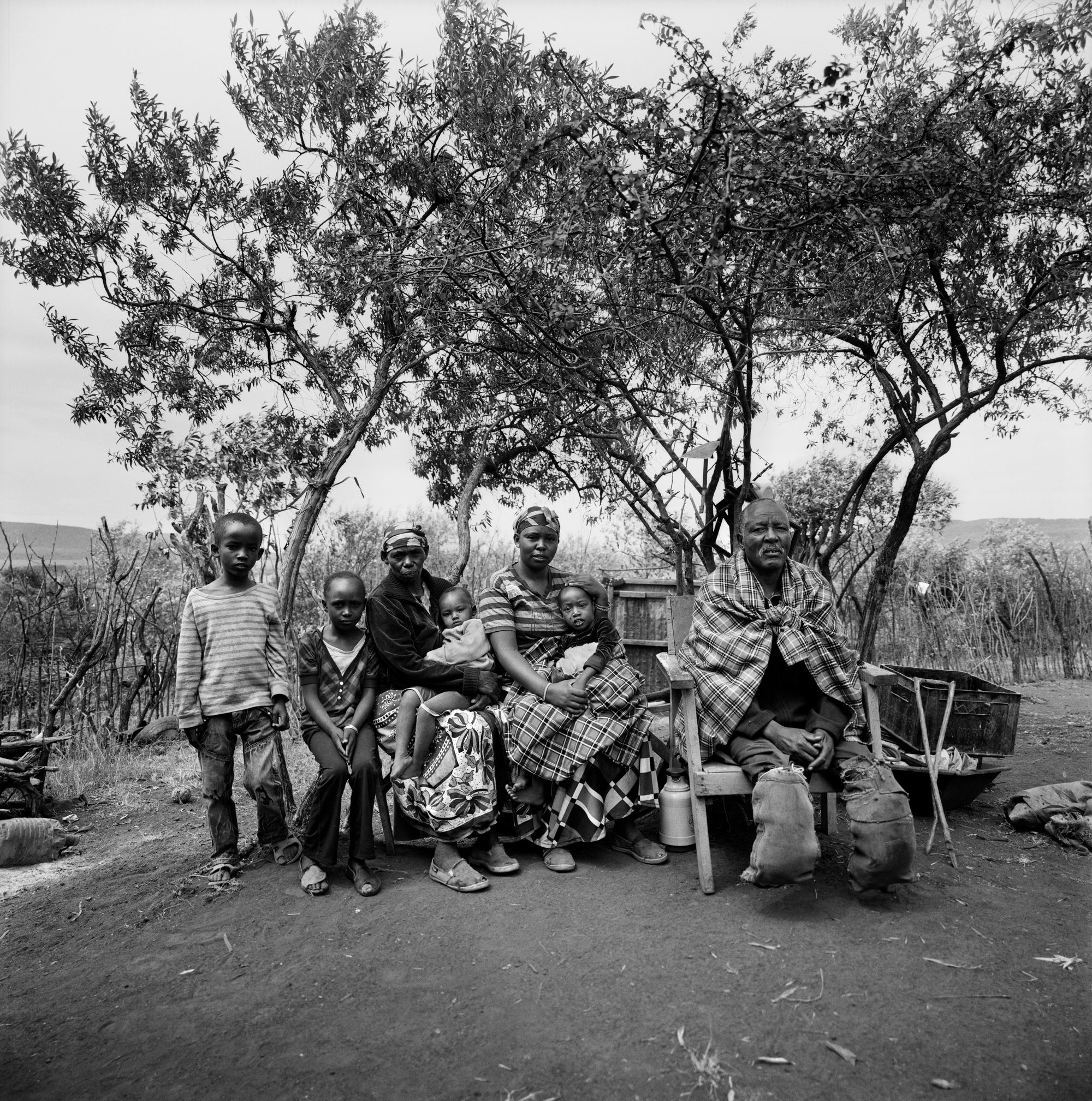Black and white photo of a family group, of a man, two women, and four children. The man appears oldest, in a situation of importance on the wooden chair, legs amputated at the feet. Next to him the two women each with a child on their lap, and two older children next to them. Behind them sparse trees, a wooden fence, and hills in the horizon.