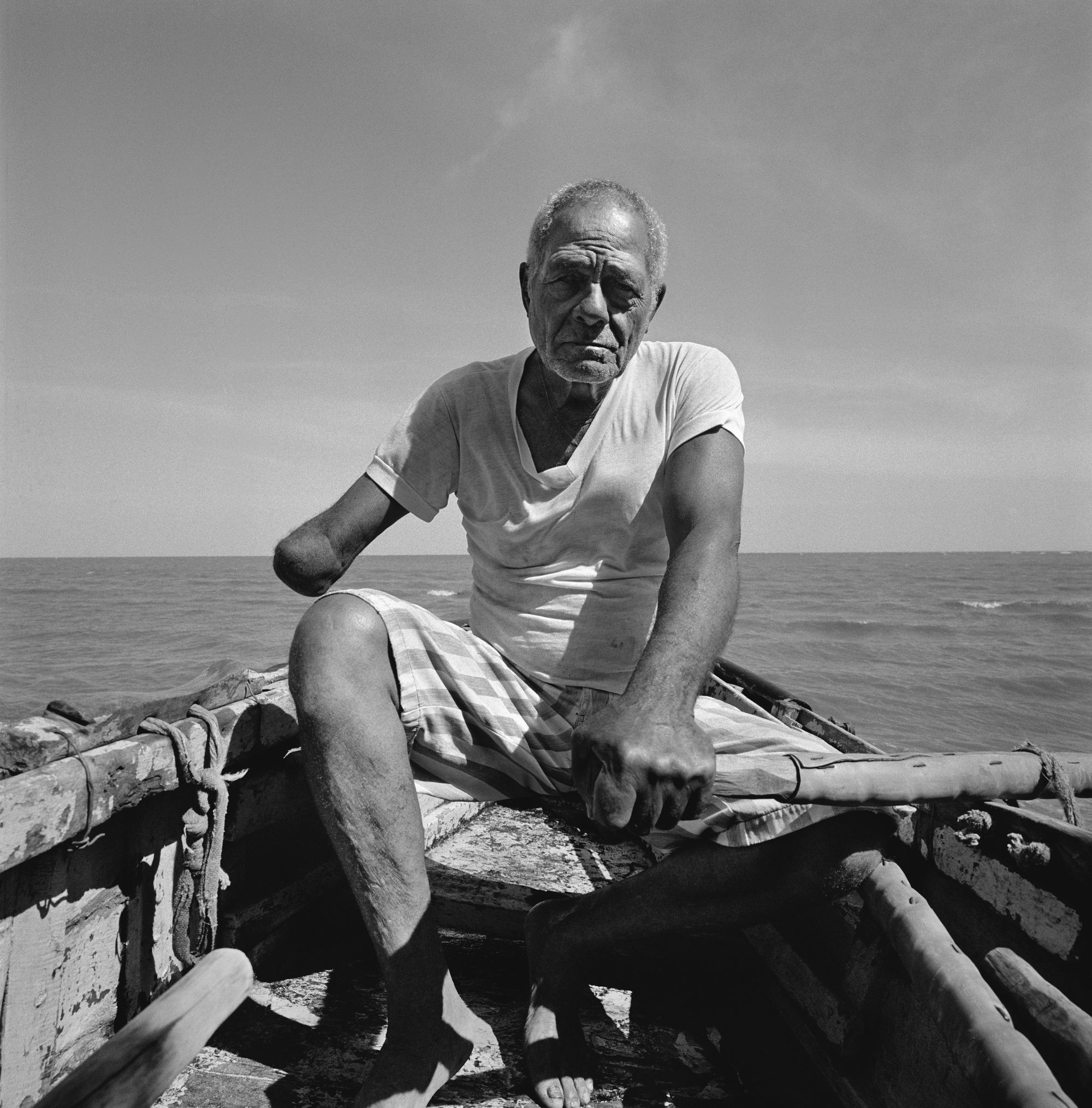 Black and white photo of an older man at sea on a rowboat. His left hand holds the handle of an oar, and his right arm is amputated. Wearing a simple vest and shorts, he looks seriously at the camera, the horizon stretching behind him.