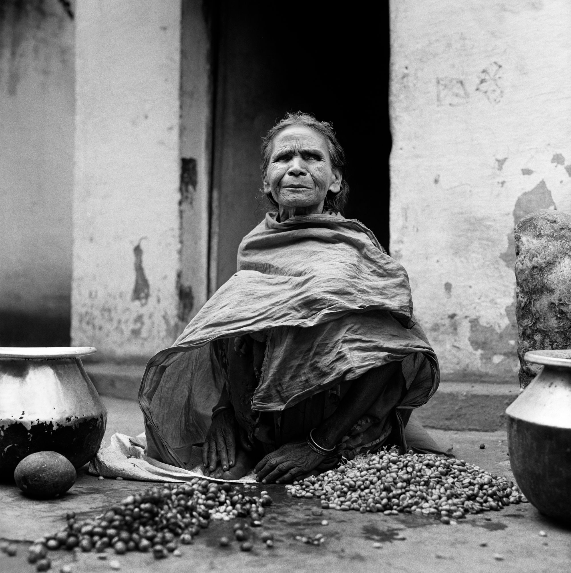 Black and white photo of an older woman, seated in front of a doorway. Her face is expressively wrinkled, perhaps with the start of a smile. Her eyes are shaded. She wears a shawl that covers her body and at her feet are piles of small fruit and large pots.