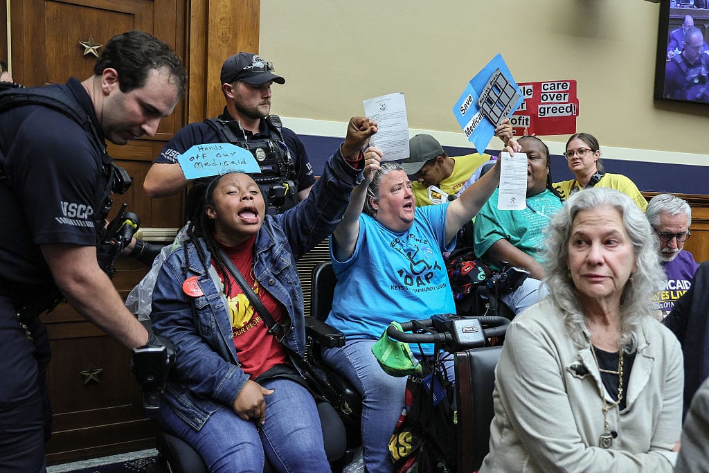 A photo of wheelchair-users protesting in a US government committee room, while two members of the USCP police intervene beside them. At the centre of the picture are three women, two of them Black and one white. They all look emotional and angry, and are shouting and raising their arms in the air, holding hand-made signs which read "Save Medicaid", "Hands Off Our Medicaid" and "People over Profit, Care Over Greed!". One of the policemen has his hands on one of the protestors' wheelchairs, as if he is trying to remove her from the room.