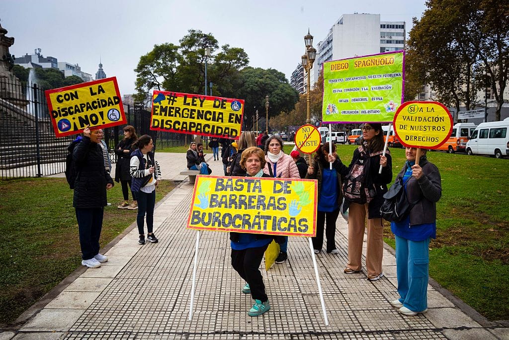 Photograph of a group of protestors holding yellow signs on a sidewalk. In the centre, a woman standing with legs bent, holds a sign reading "basta de barreras burocráticas." Other signs read "Ley de Emergencia en Discapacidad", "Con La Discapacidad, ¡No!", and "Sí a la validación de neutros derechos".