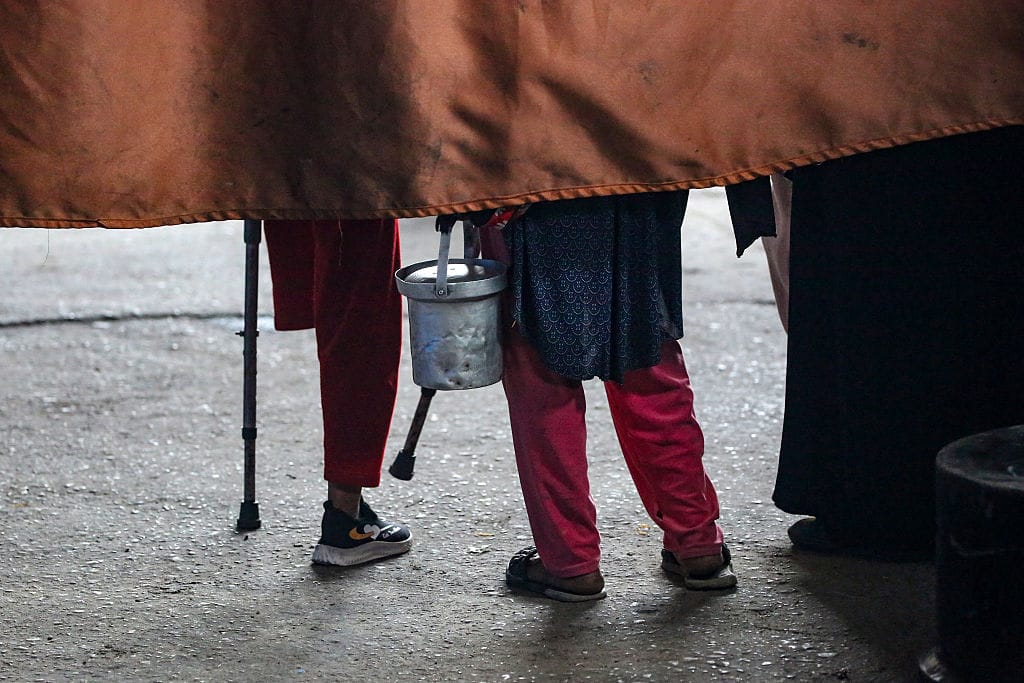 A photo of the legs of two children, one of them an above-knee amputee, as they stand behind a rusty-coloured drape. The drape hangs over the top third of the frame, so that only the backs of their legs are visible. The child who is an amputee is wearing red cropped trousers and using crutches, while the other child is wearing a blue tunic and pink trousers, and is holding an aluminium saucepan.