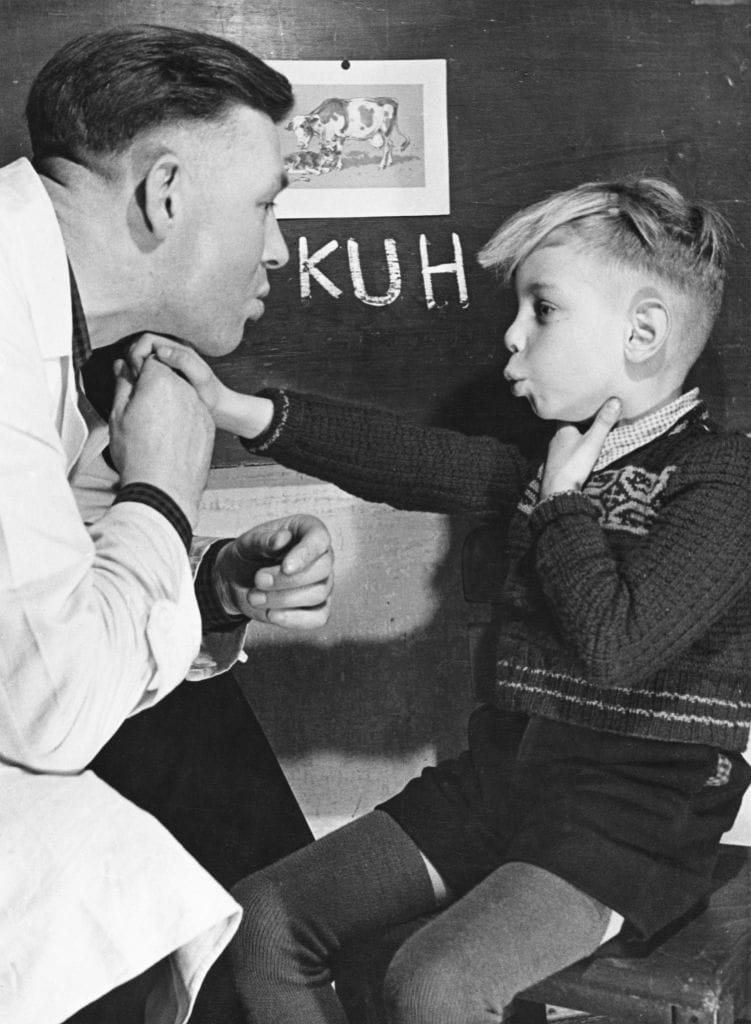 An old black-and-white-photo shows an instructor with a long white coat leaning over a boy, as they both mouth a word. The boy has one hand on his own throat and the other held against his instructor's. Behind them, on a blackboard, the word "Kuh" is written in chalk alongside an illustration of a cow. The boy has short blonde hair, wears a jumper with shorts and winter leggings. 