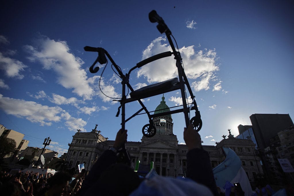 A photo of a walker being held in the air in front of the Argentine National Congress. The walker is dark silhouette against the sky, which is bright blue with a few clouds. Underneath the walker are banners or flags, in Argentine colours, and a crowd of people with hands in the air, as if in celebration.