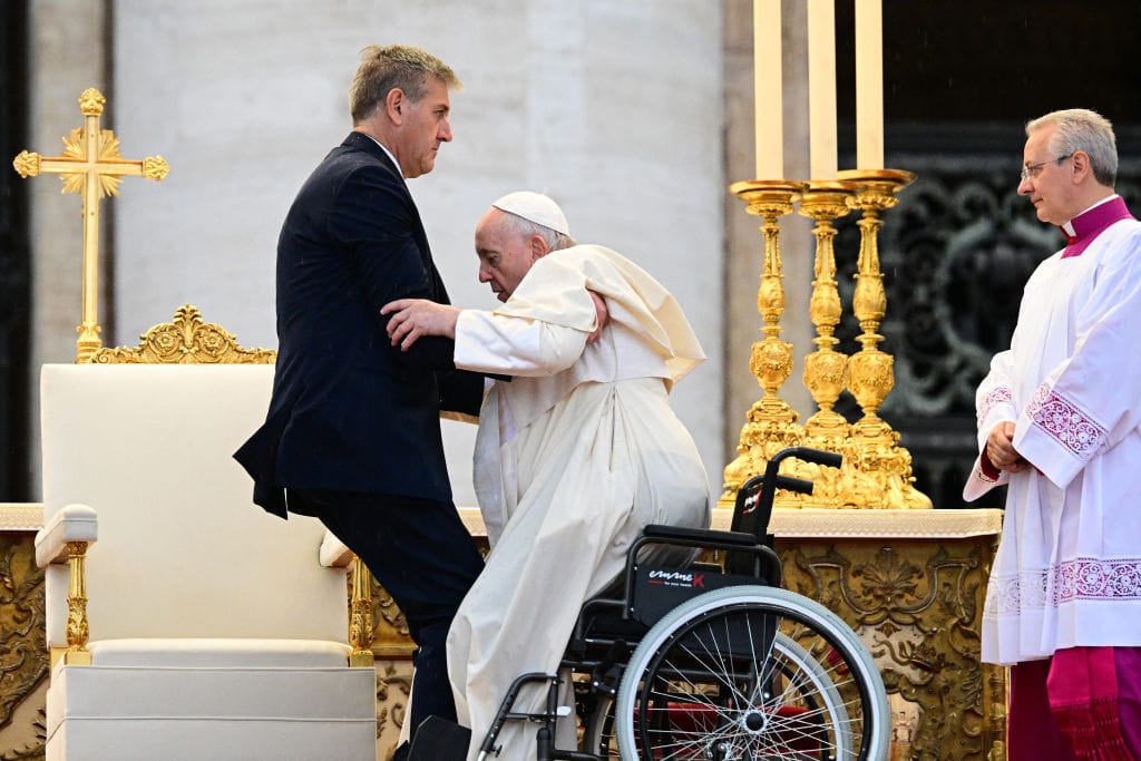 A photograph of Pope Francis being lifted out of his wheelchair by a tall man wearing a dark suit. The Pope is wearing a hat and white robe that bunches as he is lifted. They are on stage with ornate golden furniture, next to a large white chair. Behind them there are huge candlesticks and a cross. A third man, in white and purple robes, looks on with a slight smile.