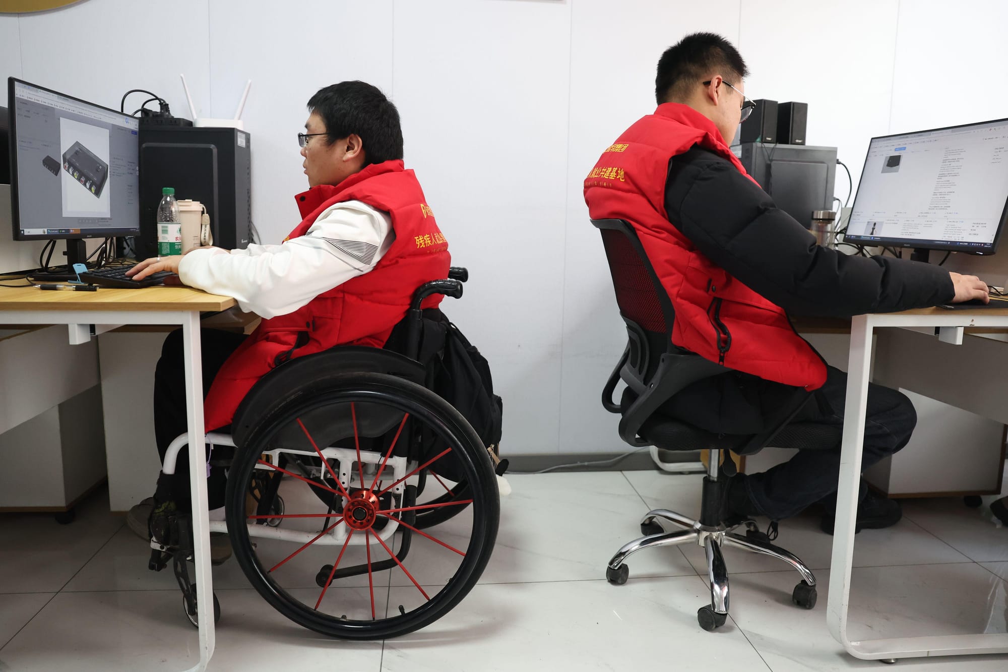 A photo, taken in profile, of two Chinese people, one of whom is a wheelchair-user, sat in front of computers at their two desks in an office. They are both wearing glasses, matching bright red gilets with yellow branding, and black trousers, and they are concentrating on their computer screens and typing at their keyboards. On the screen on the left, which belongs to the wheelchair-user, is a large photo of some kind of black electrical port.