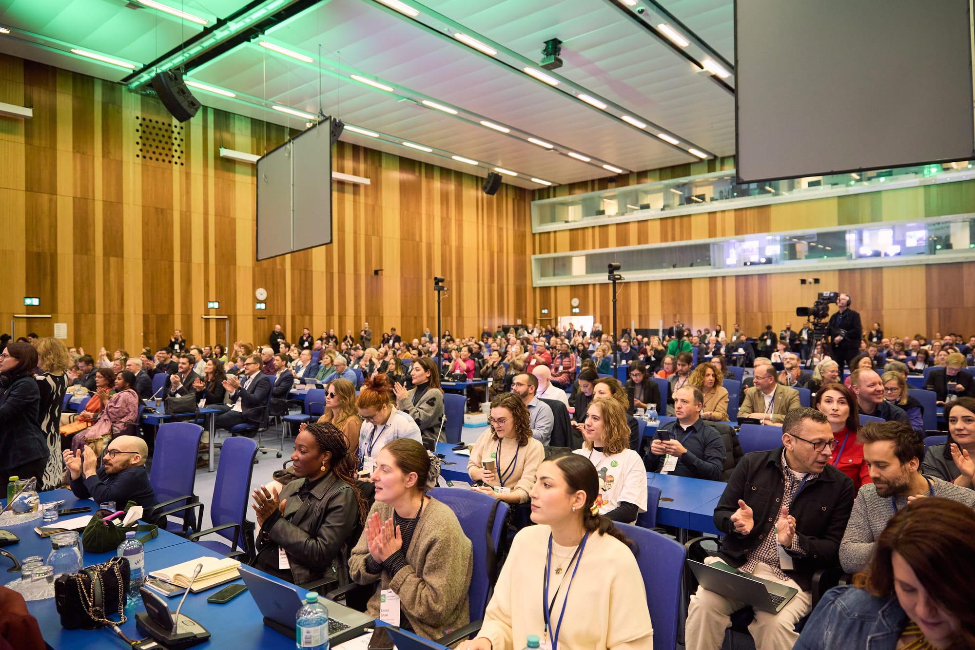A photo of the audience in a large conference room with rows of people seated. Many are smiling or applauding, some are simultaneously gossiping. The conference room has wood panelled walls and many of the audience are also using their phones or laptops. There are screens and cameramen visible. 