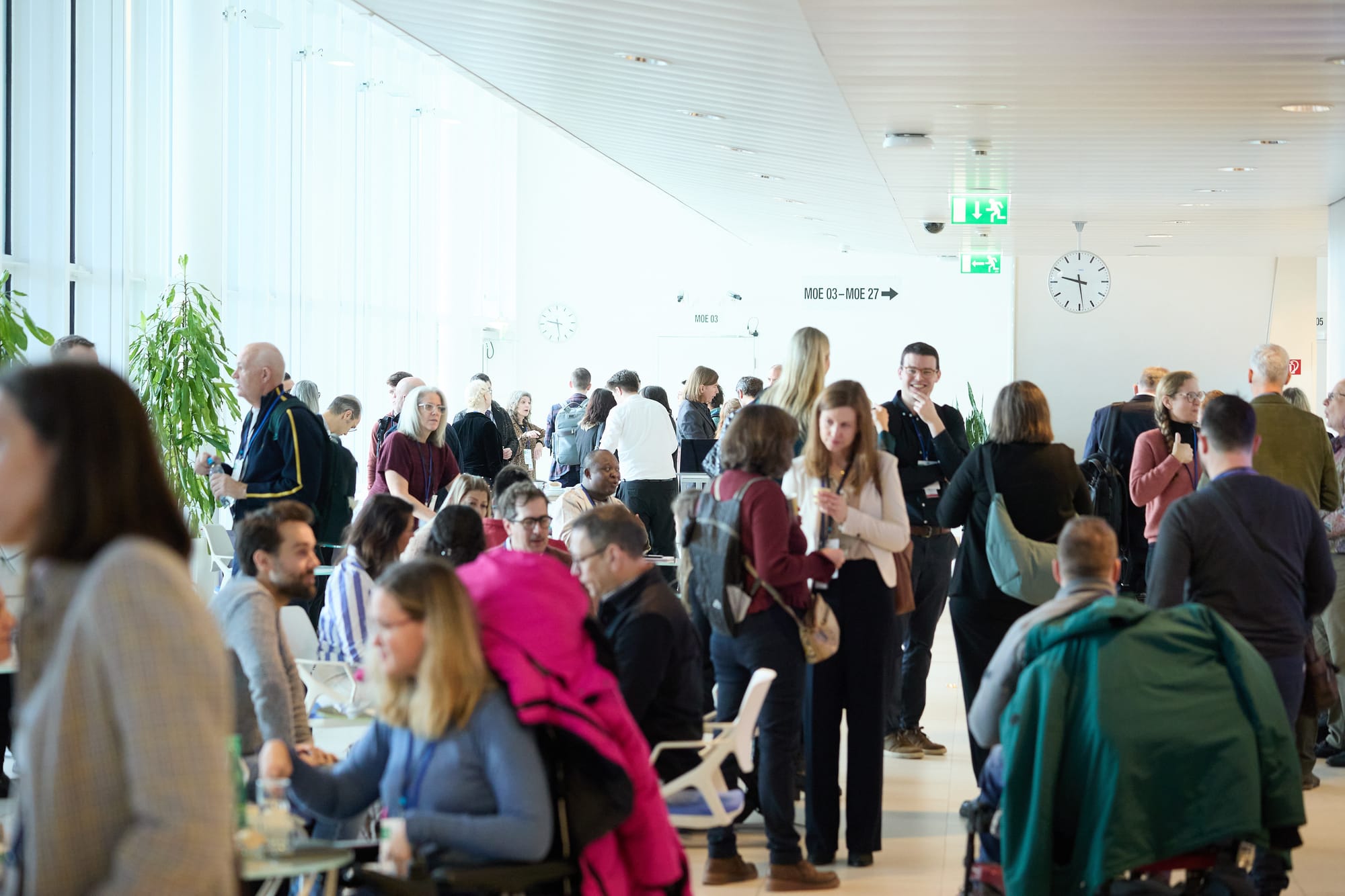 A photo of a busy, large, light-filled room packed with small groups of people chatting animatedly. The people are mostly talking in pairs or in groups of three's, and some of them are seated at tables, while others are sitting in their wheelchairs or standing. They are a mix of ages and ethnicities, although the majority seem to be white. 