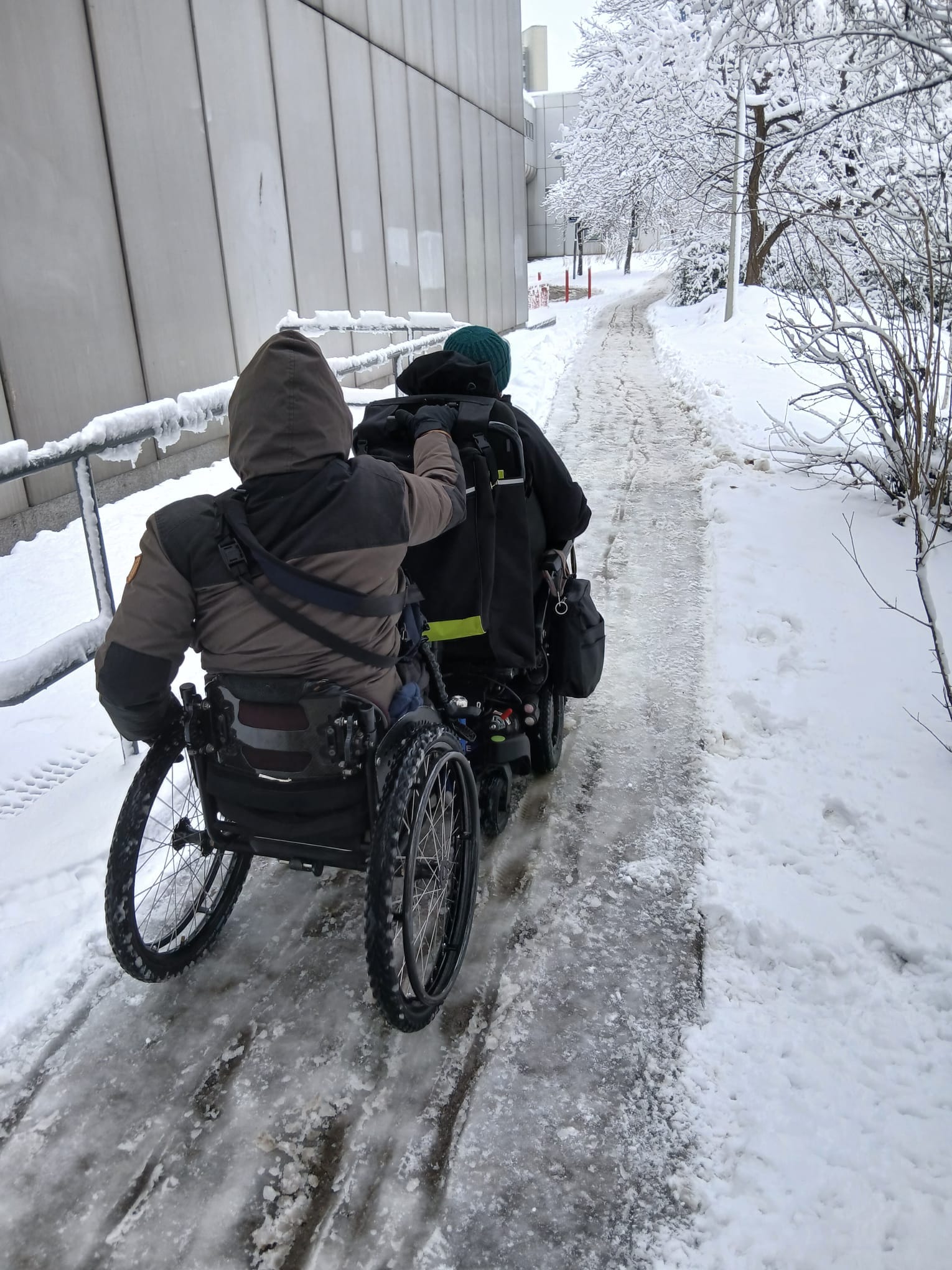 A photo of two male wheelchair-users going up an icy path through the snow. Behind a man uses a manual wheelchair and he holds onto the back of the electric wheelchair in front. Both wear winter clothes, and there are trees along one side of the path.