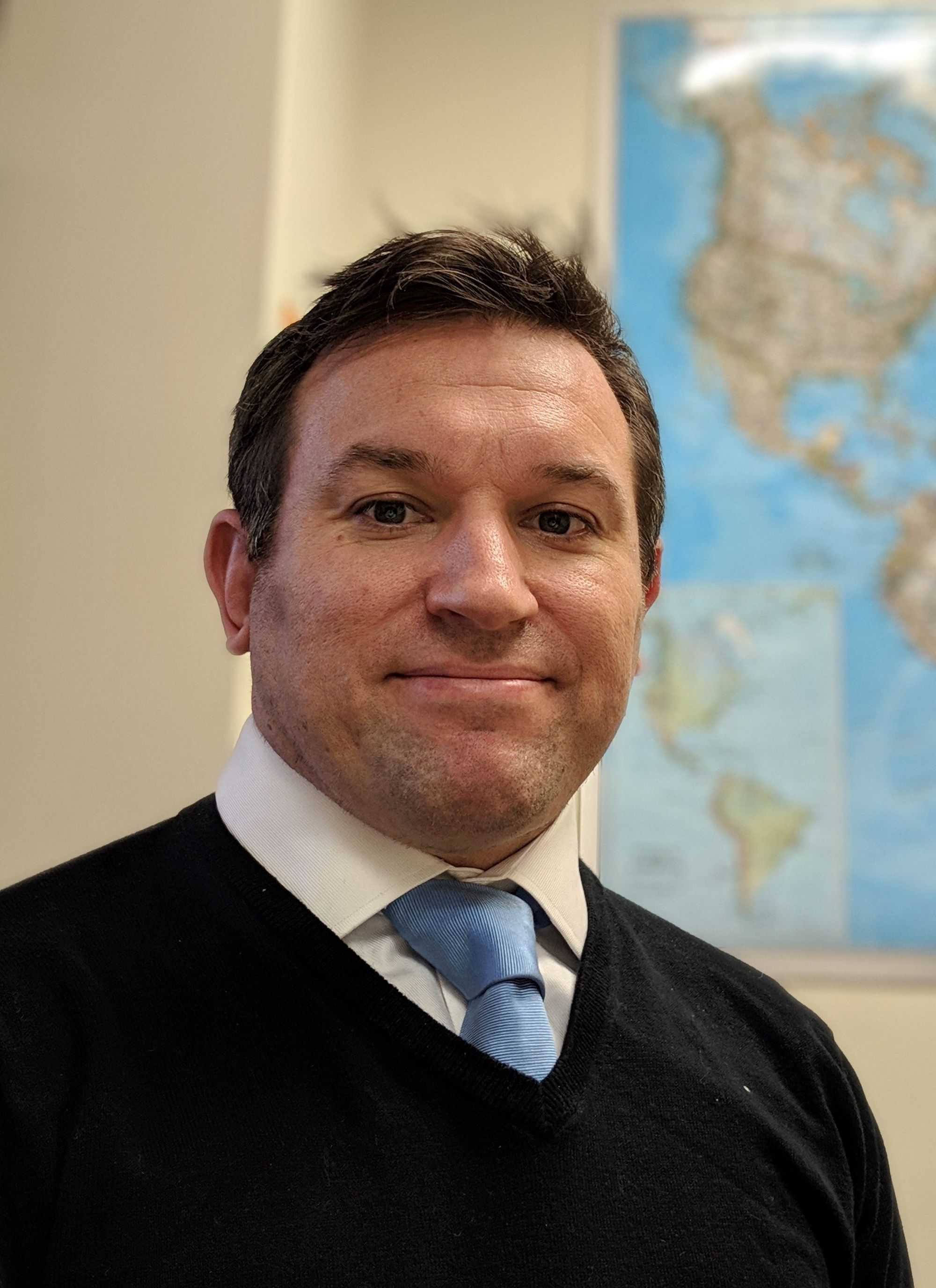 Profile of Antony looking forward with slight smile. White man with short dark hair in front of a world map. He wears a blue tie and black sweater. Profile of Antony looking forward with slight smile. White man with short dark hair in front of a world map. He wears a blue tie and black sweater.