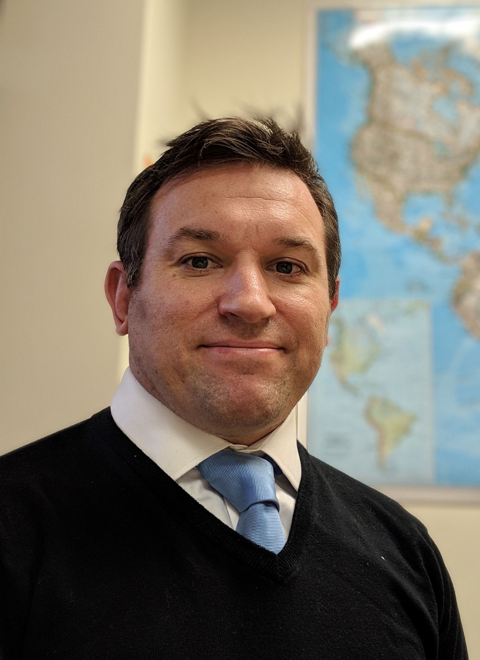 Profile of Antony looking forward with slight smile. White man with short dark hair in front of a world map. He wears a blue tie and black sweater.