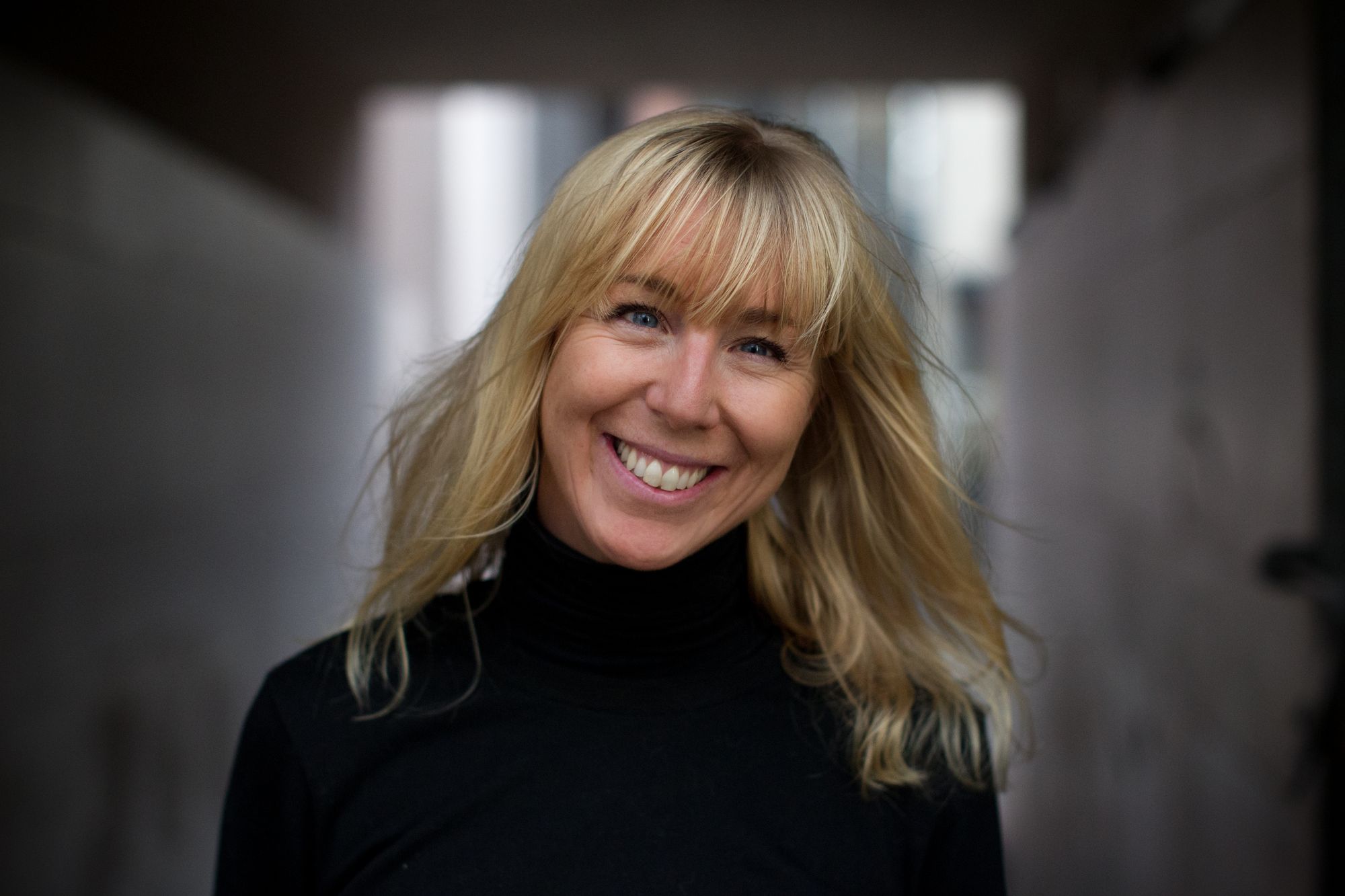 Caroline with an open smile to camera, a white woman with blonde hair to her shoulders, wearing a black top and against a blurred background.