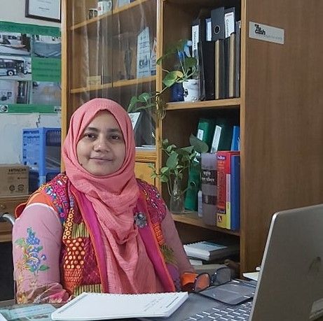 Misti sits in her office, looking towards camera she wears a pink headscarf and a colourful pink top. Her laptop frames the foreground and a filing cabinet with files and plants the background. 