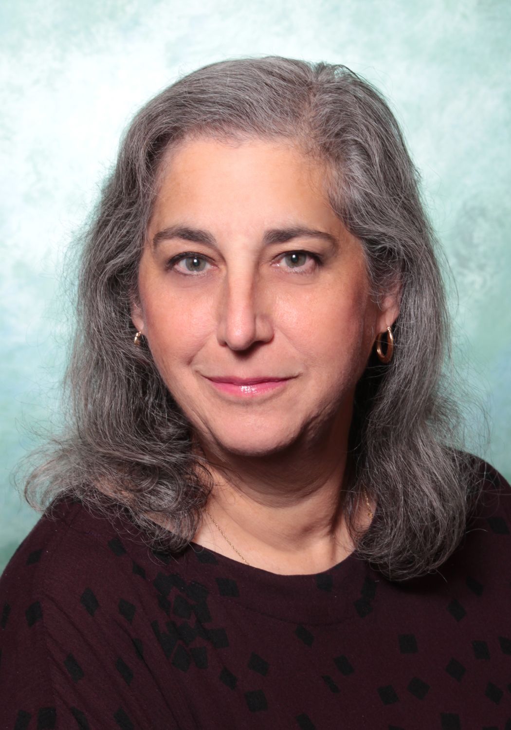 Head-shot of Jennifer, a white woman with grey-black hair, against a pale studio background. Jennifer looks inquisitively to camera, with a dark red top and a small smile brought out by pink lipstick. 