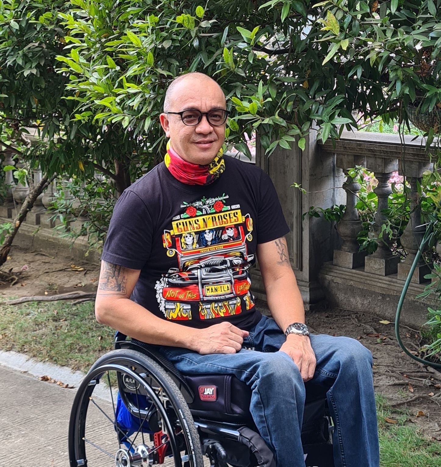 A photograph of Abner, a Phllippinian man, sitting in a wheelchair, half-smiling in a knowing way towards camera. Abner has dark circular glasses and is wearing jeans with a black Guns N’ Roses Manila t-shirt and a neck-scarf in red and yellow colours matching the t-shirt illustration. He has tattoos on his upper arms, and the background is green vegetation with an elegant stone wall. A photograph of Abner, a Phllippinian man, sitting in a wheelchair, half-smiling in a knowing way towards camera. Abner has dark circular glasses and is wearing jeans with a black Guns N’ Roses Manila t-shirt and a neck-scarf in red and yellow colours matching the t-shirt illustration. He has tattoos on his upper arms, and the background is green vegetation with an elegant stone wall.