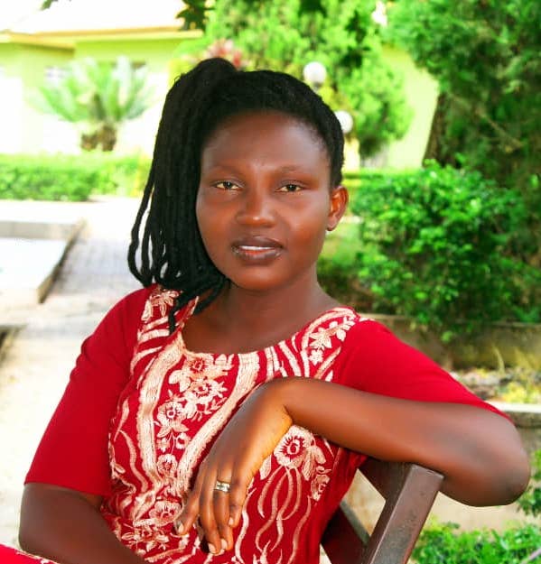 Bernice sits, with one arm on the back of her chair, looking towards the camera. She is a black woman wearing a red dress with embroidered patterns. She has black hair braided in a ponytail. 