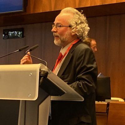 Stefan at a podium with microphones - Stefan is white, brushed-back white hair and a beard, glasses, wearing a suit, making a point with his right hand