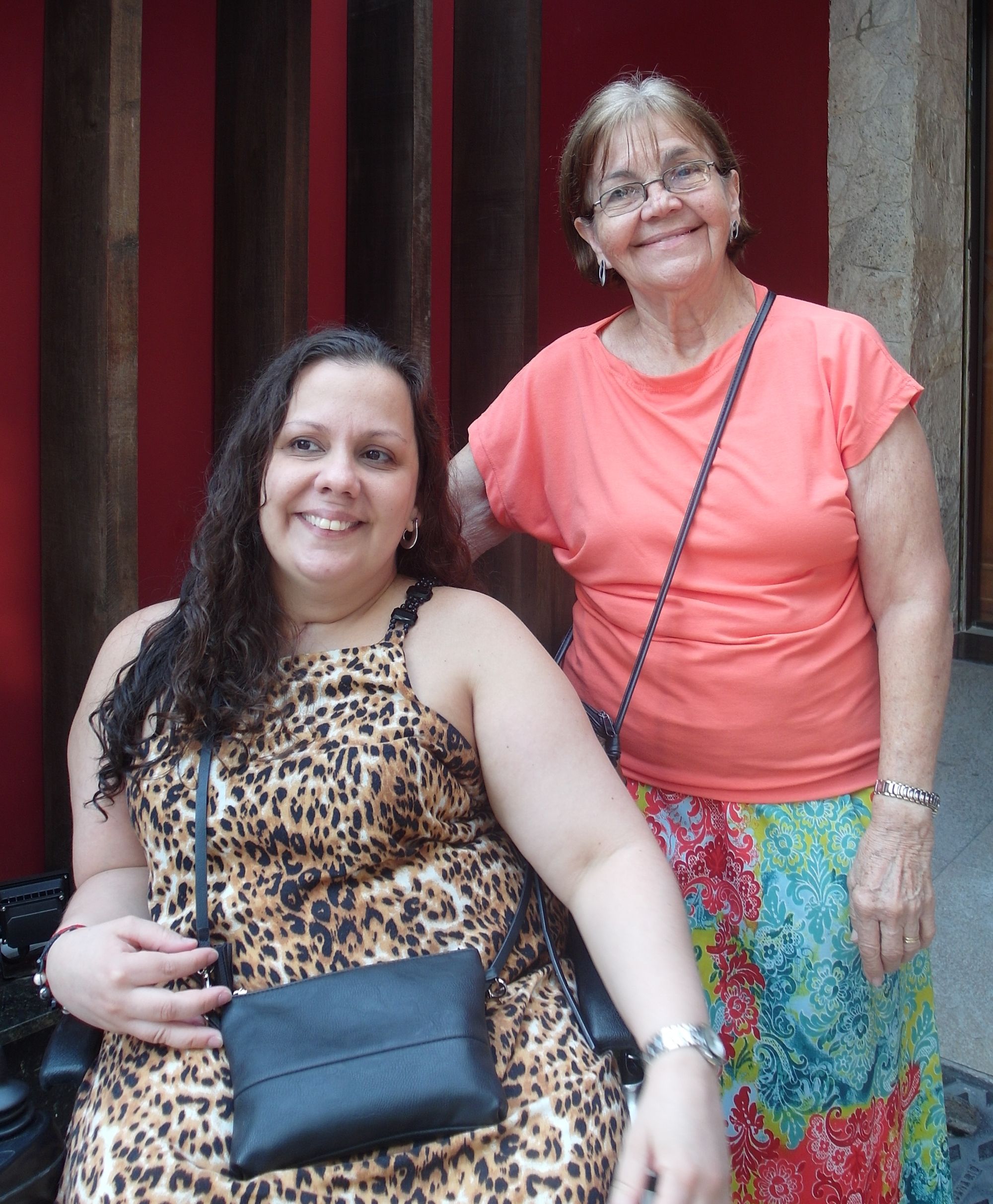 Sheila smiles with a glance off camera, dark hair curling down, sitting in a wheelchair with a leopard-print dress. Raylda to her right is an older woman smiling towards the camera and rose coloured top and colourful print dress.