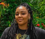 Profile picture of Tharā, a multiracial woman with dreadlocks. Tharā wears big earrings and a black t-shirt. In the background is green vegetation.