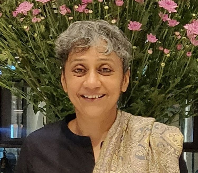 A profile picture of brown-skinned woman almost smiling. Wearing a black top with a gold shawl she has short tousled hair.