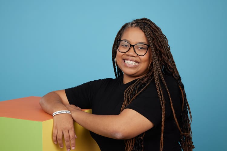 A photo of Luciana, a young Black woman wearing a black t-shirt, oversized dark-rimmed glasses and her hair in long braids.