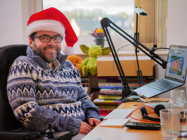A photo of Peter, a white man with a dark beard, blue eyes and dark-framed glasses, wearing a Santa hat and smiling at the camera.