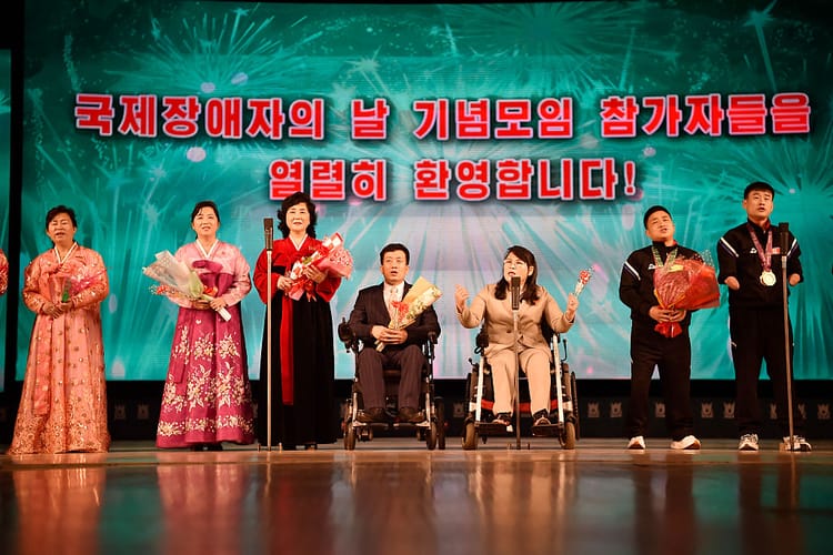 A brightly coloured photograph of seven men and women, several of whom are wheelchair-users or visibly disabled. They are side by side on a kitsch, carefully choreographed stage.