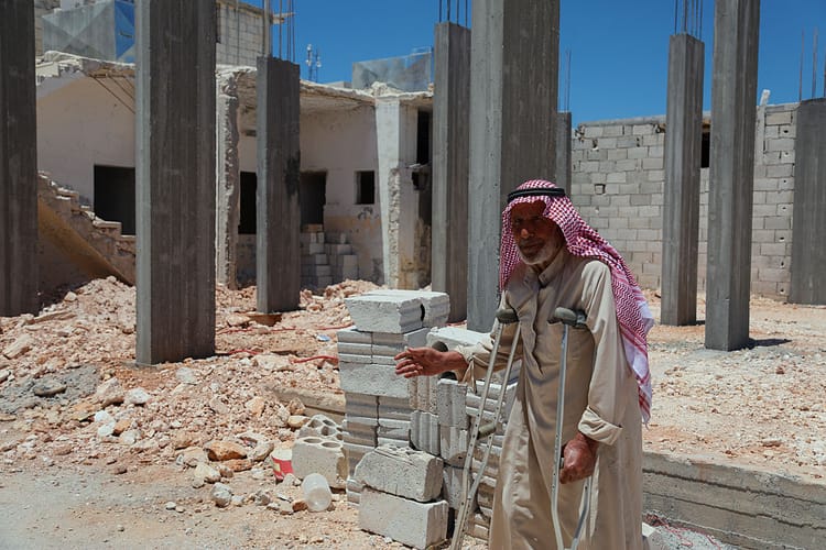 A photo of a late middle-aged man supporting himself on crutches and wearing a red keffiyeh as he stands in front of a construction site.