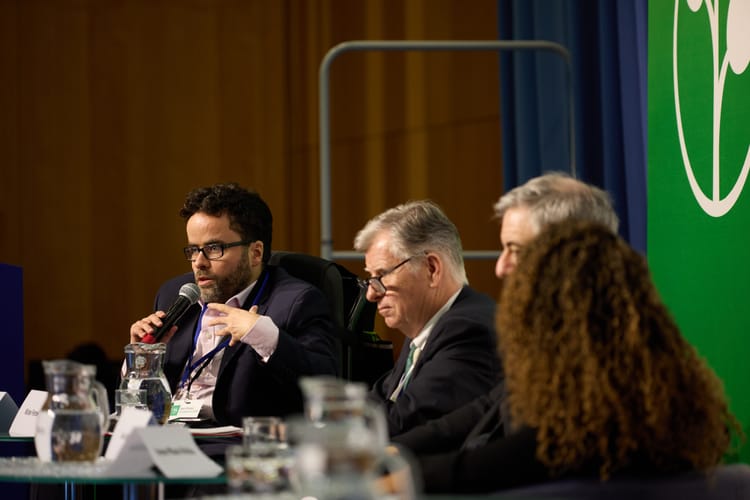 A photo of Peter, a white man with dark hair, beard and glasses, wearing a pink shirt and navy blue suit and talking into a microphone as he sits beside three other people on a panel.