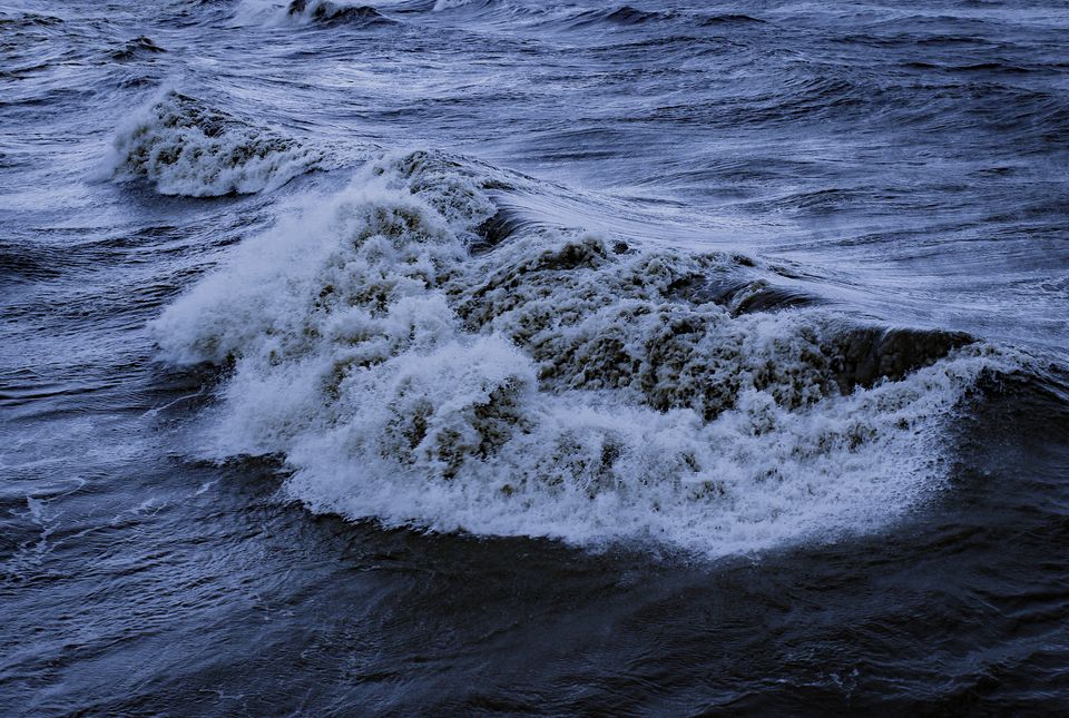 Photograph of waves crashing on the shore. The sea is dark grey blue and the waves froth white.